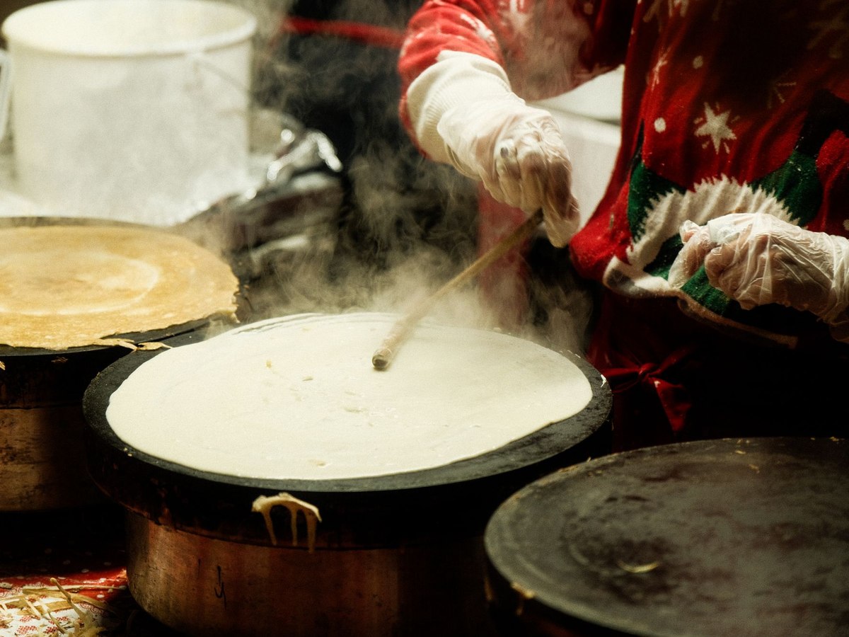 Pâte à crêpes étalée sur une crêpière fumante, travaillée par des mains gantées