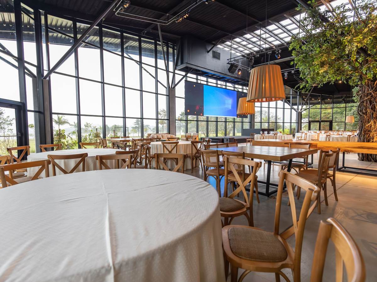 Salle de restaurant vide avec des tables rondes nappées de blanc et des chaises en bois sous une grande verrière
