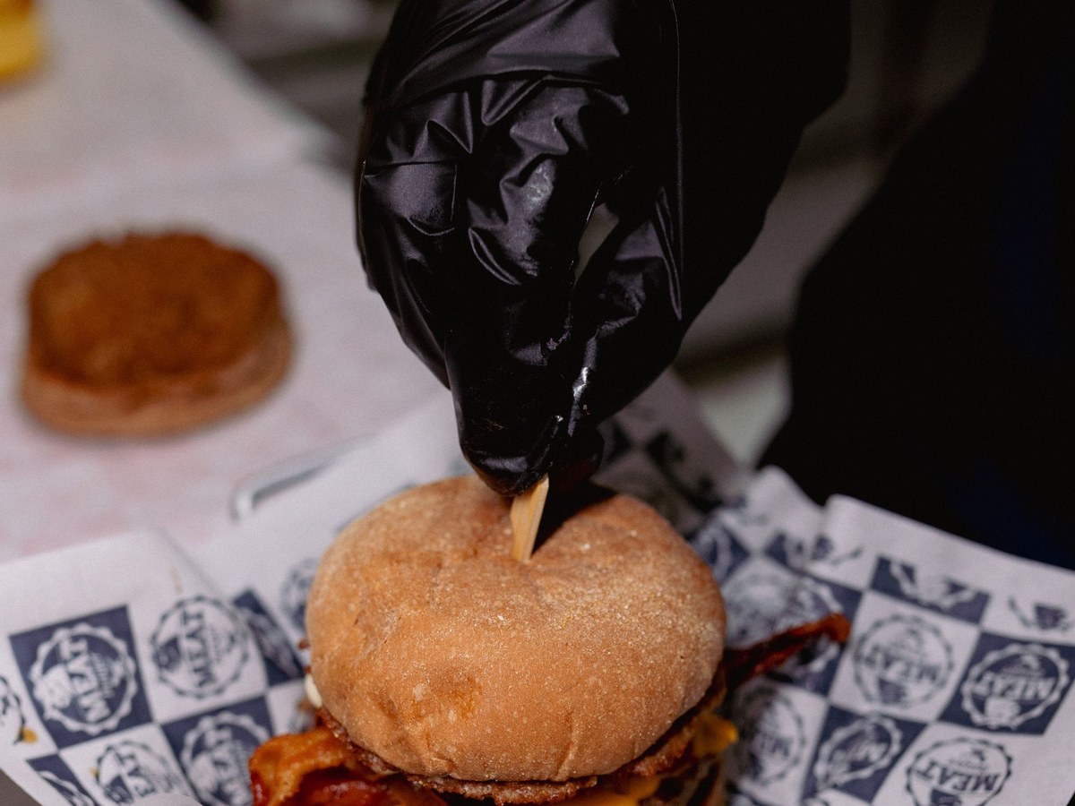 Burger en préparation : une main gantée de noir plante une pique en bois dans le pain, sur du papier d'emballage